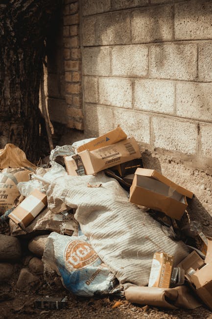 A pile of mixed waste materials located at the base of a brick and concrete wall, consisting of flattened cardboard boxes, some with visible printed labels and barcode stickers, partially covered by a textured, off-white fabric sack. The cardboard appears in various shades of brown with some torn edges, indicating that they have been recently discarded. Surrounding the waste are small rocks and dirt, with a dark tree trunk partially visible on the left side of the image. The background features an aged brick section on the left and a concrete block wall on the right, both showing signs of weathering. The scene suggests an outdoor or semi-enclosed area potentially prepared for private rubbish collection or on-site clearance, as managed by local waste removal services such as Rubbish Clearance Roehampton. The overall lighting is natural, with soft ambient light highlighting the textures of the materials and surfaces, indicating a typical daytime setting for waste disposal activity relevant to private rubbish removal and waste management solutions.