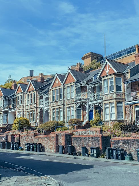 A row of Victorian-style terraced houses with decorative white woodwork and bay windows, situated along a residential street. The buildings are constructed with red brick and feature pitched roofs with grey tiles, some with small front gardens enclosed by low brick walls. In the foreground, black wheelie bins are lined up along the pavement, indicating waste collection points. The street is quiet, with no visible pedestrians or vehicles, and the sky above is clear with a few wispy clouds, suggesting a bright, sunny day. The overall scene reflects a typical urban residential area where private waste disposal services, such as those provided by Rubbish Clearance Roehampton, may handle rubbish removal for the properties. The environment is tidy and well-maintained, with greenery in the form of bushes and small trees in front gardens, and the scene emphasizes the importance of organized waste management in urban living spaces.