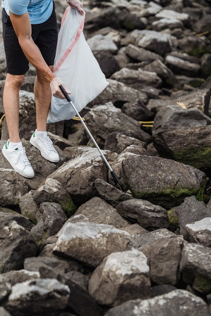 A person wearing white sneakers, black shorts, and a blue t-shirt is using a grabber tool to pick up litter from a rocky shoreline. The individual is holding a large white trash bag with their other hand, which appears to be partially filled, indicating an effort to clear debris from the area. The rocks vary in size and color, ranging from dark gray to brown, with some showing patches of moss or algae, characteristic of a coastal environment. The scene is outdoors with natural lighting, and the rocks are scattered across the ground, forming an uneven surface typical of a rugged shoreline. This visual highlights an active waste collection effort in a natural setting, aligning with private rubbish removal or on-site clearance services like those provided by Rubbish Clearance Roehampton, which facilitate environmentally responsible waste disposal outside traditional municipal collection methods.